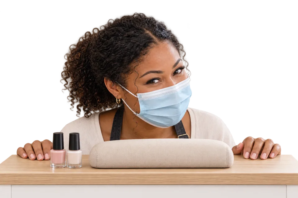 A nail technician wearing a mask and peeking over a manicure desk