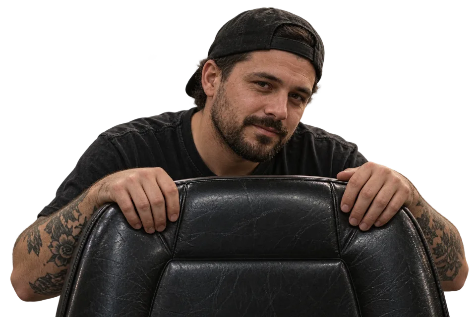 A tattooed artist in a backwards cap peeking over a worn black tattoo chair